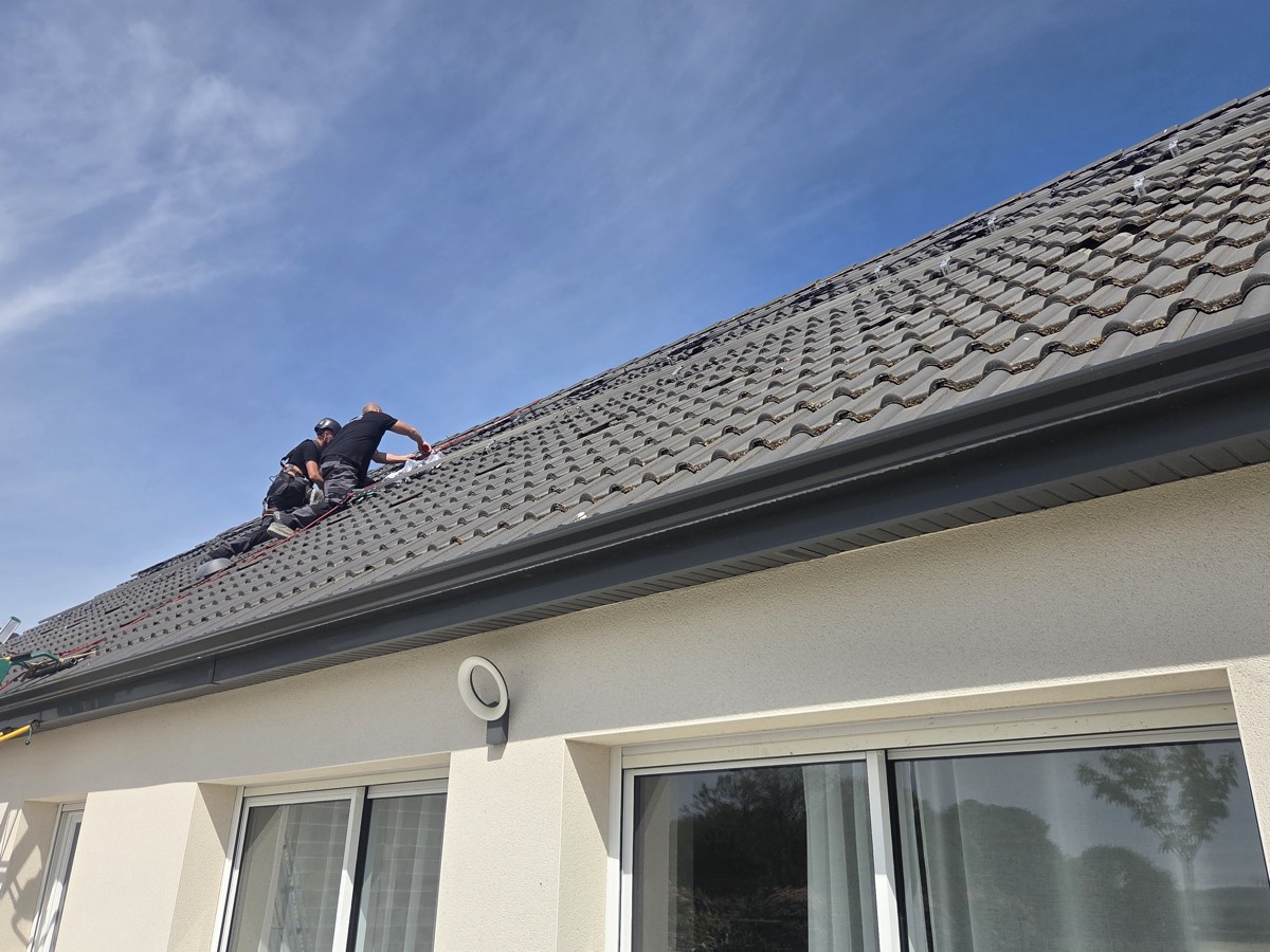 Deux installateurs travaillent sur le rampant d'une toiture, tuiles déposées.