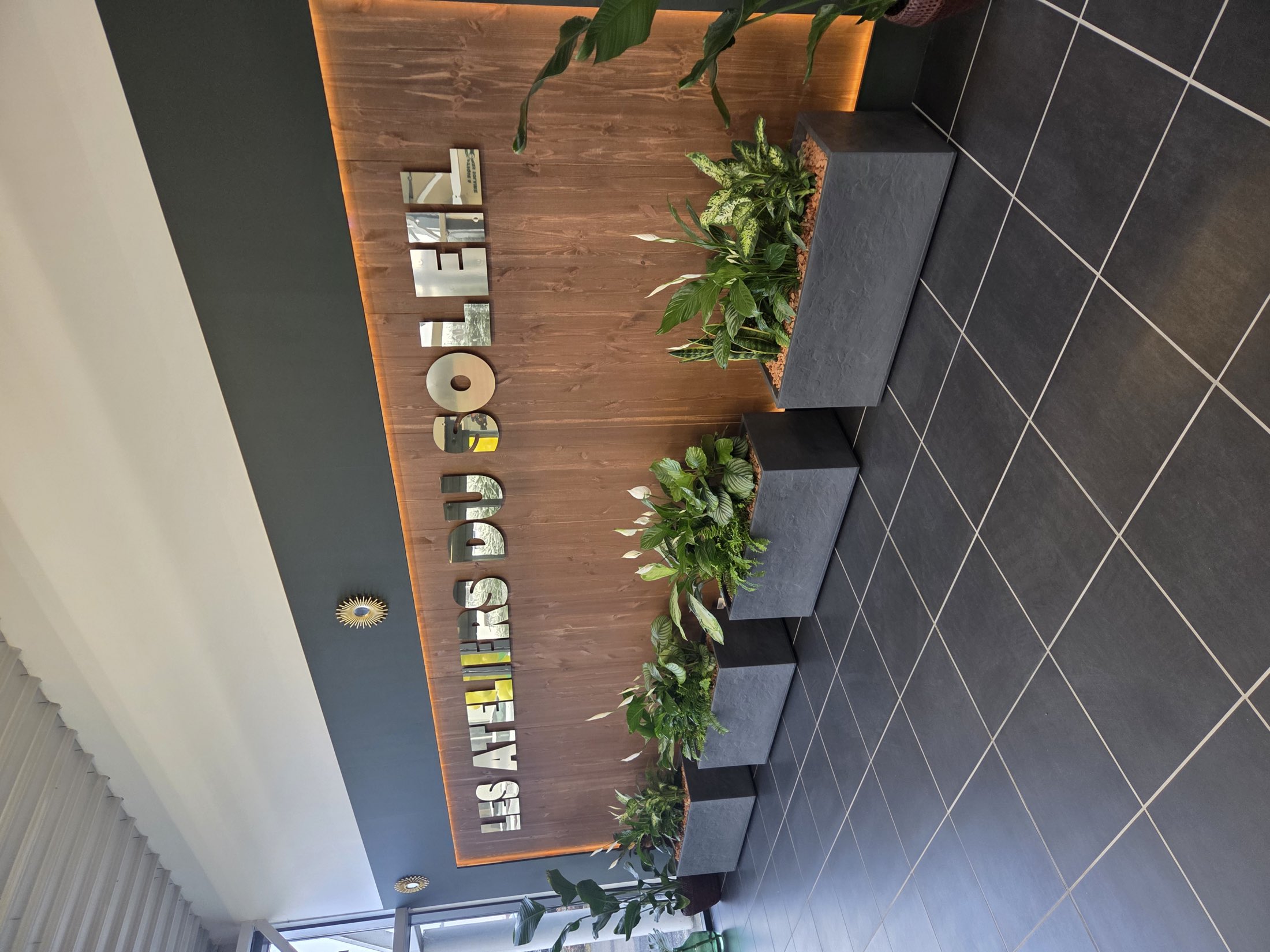 Le mur d'entrée du showroom, avec le nom Les Ateliers du Soleil en lettres découpées et une rangée de plantes.